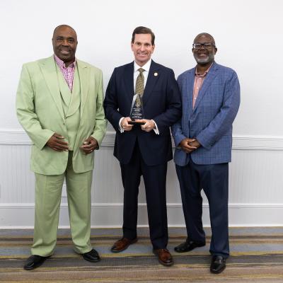 Three men are standing together in a formal setting, with the man in the center holding an award.
