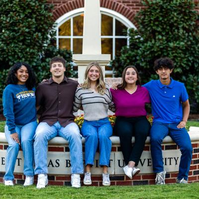 Five college students sit on a circular raised flower bed surrounding ETBU's Light on the Hill