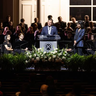 Many people on a stage in a dark lit room with a light fixed on a speaker stood at a podium