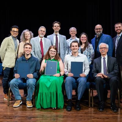 A group of people sitting and standing on a dimly lit stage smiling for a group photo