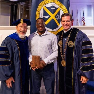 3 people stood in front of the seal for East Texas Baptist University, in a well-lit room wearing academic regalia