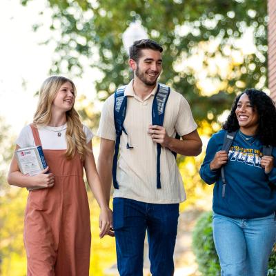 Three college students walk and talk together on campus, smiling and carrying backpacks and books.