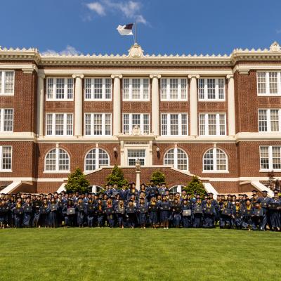 A large group of graduates in graduation regalia stand in front of ETBU's Historic Marshall Hall