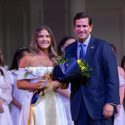 2 people in formal clothing smiling in front of a large group of people in white clothing behind them