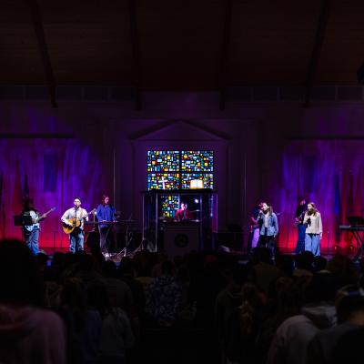 6 people standing on a dimly lit stage holding instruments in front of a large auditorium crowd