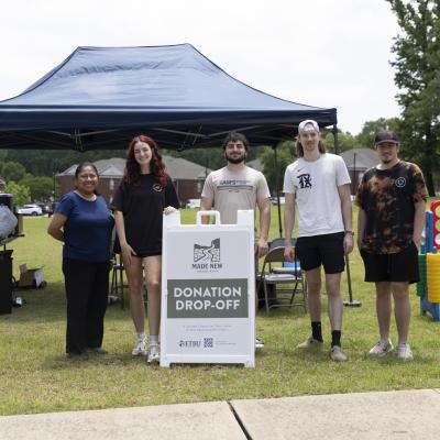 Five people stand in front of a blue canopy tent at a donation drop-off event, with a sign reading "Donation Drop-Off" placed in the center.