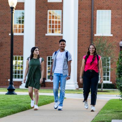 Two females and one male ETBU student walking down a side walk in front of a red brick building with white columns in the background.