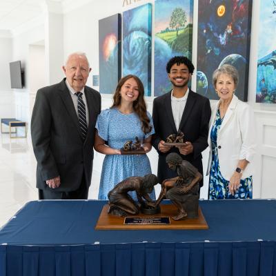 Bob and Gayle Riley stand on either side of a male and female student holding awards behind a table inside a building with white walls.