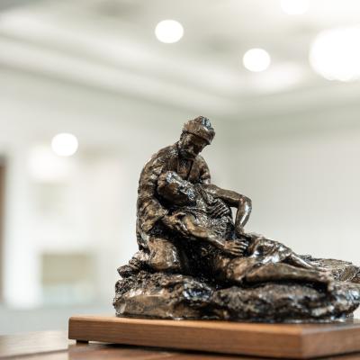Bronze sculpture of the Good Samaritan kneeling and cradling an injured man, displayed on a wooden base in a softly lit indoor space.