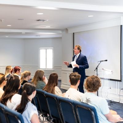 Rows of students are seated in blue chairs listening to a speaker who is standing in front of a projector screen.