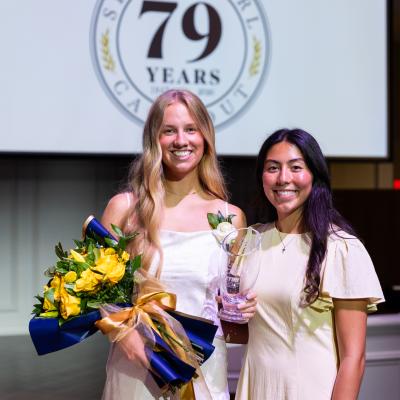 Two girls stand in white dresses. One is holding a bouquet of flowers and there is an event slide shown on a projector screen behind them.