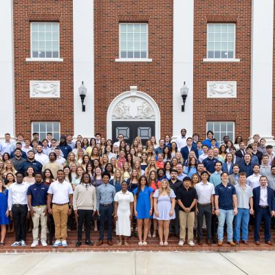 A large group of more than 100 students stands on an exterior staircase showing their class rings