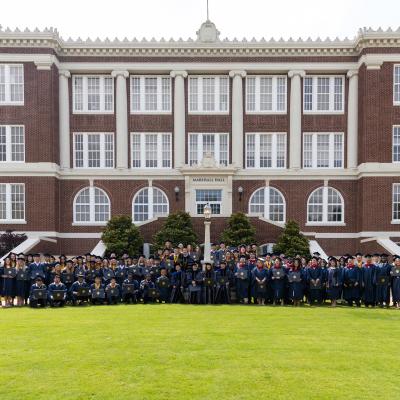 A group of graduates in navy regalia standing in front of a red brick building smiling.