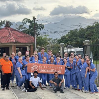 A group of students in blue scrubs stand with a International Medical Relief orange sign in front of a scenic backdrop.