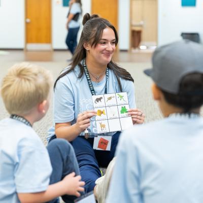 Female sitting on the ground with a group of young children holds a sound chart up while smiling. All are wearing light blue shirts.