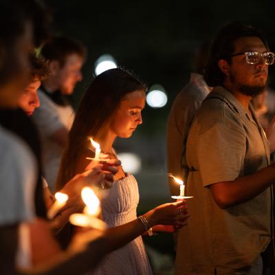 Photograph of a candlelight vigil held outdoors at night, showing a group of people holding lit candles with solemn expressions. Warm candlelight illuminates faces against a dark background, highlighting a sense of unity and remembrance.