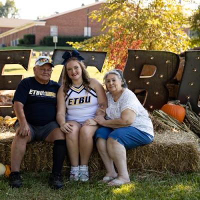 Female student sitting on hay bale with her parents smiling at camera