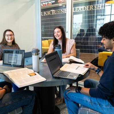 Two male and two female students sitting at a circular table with books and laptops smiling and laughing.