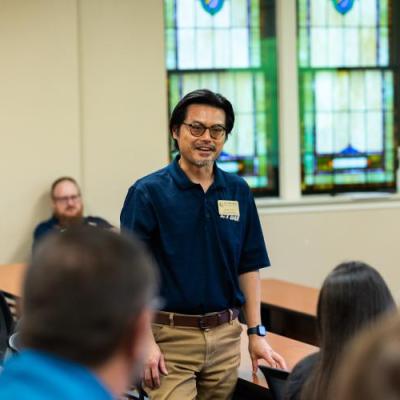 A man in a navy shirt stands and smiles while speaking to a seated group in a classroom with stained glass windows.
