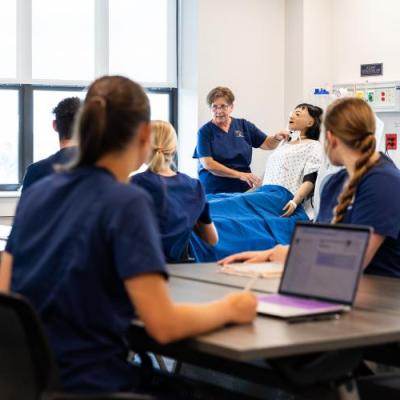 Medical instructor demonstrating a procedure on a mannequin in a classroom setting with students observing.