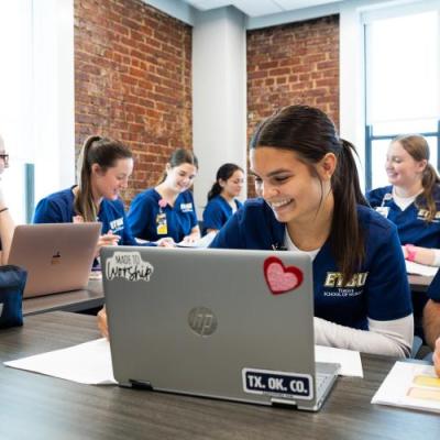 Students in navy scrubs work together on laptops and notes in a classroom setting.