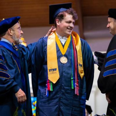 Two professors in academic regalia place a stole around a male graduate on a stage.