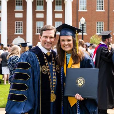 A man in navy academic regalia and a female in a navy cap and gown holding a degree smile in front of a red brick building.