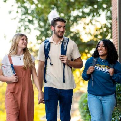 Three college students walking and smiling together on a campus pathway surrounded by greenery and brick buildings.
