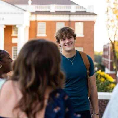 A group of male and female students talking holding backpacks and books outside on a sunny day.