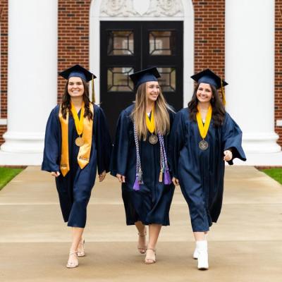 Three graduates in navy regalia walking in front of a red brick building with white columns.