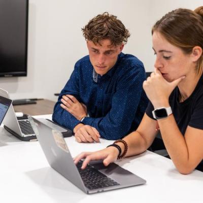 2 people sitting at a table in a brightly lit room looking at laptops