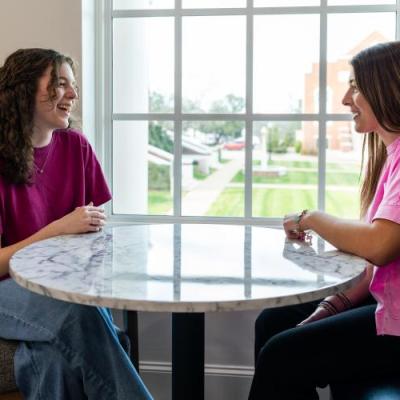 2 people inside seated at a table talking in front of a window