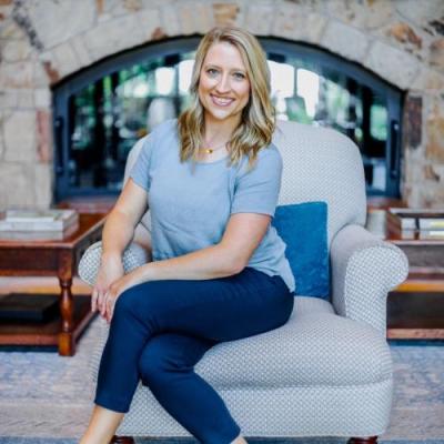 A woman in a blue shirt and jeans sitting in a grey chair in front of stone fireplace.