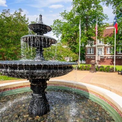 Water fountain in the foreground with flag poles in the back right on a sunny blue-skied day.
