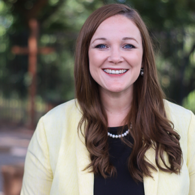 Headshot of a woman in a blazer and black shirt smiling in front of green trees.