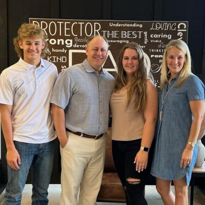 A family photo featuring four people standing side by side, smiling at the camera. From left to right: a young man in a white polo shirt and jeans with curly hair, an older man in a light gray short-sleeved button-up shirt and beige pants, a young woman in a beige sleeveless top and black ripped jeans with long hair, and a woman in a denim dress with blond hair. Behind them is a brown sign with various positive words, including "PROTECTOR," "STRONG," "THE BEST," and "LOVING."