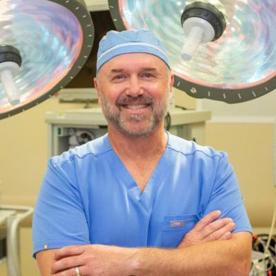 A man in blue medical scrubs, arms crossed, standing in a surgical room with overhead lights.