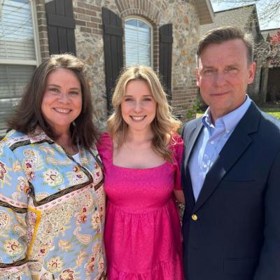 This image shows three people standing together in front of a brick and stone house with arched windows and dark wooden shutters. The woman on the left is wearing a light blue patterned blouse with white pants. The young woman in the center is wearing a bright pink dress with short sleeves and a square neckline. The man on the right is dressed in a dark navy blazer, light blue dress shirt, and jeans. All three are smiling and standing close together, posing for the photo in a sunny outdoor setting.