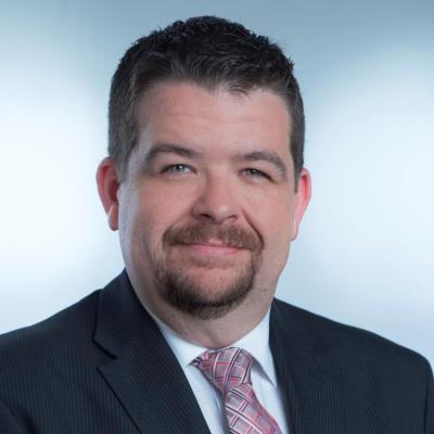 Man with short dark hair and goatee, wearing a dark suit, white shirt, and light patterned tie, with a neutral expression.