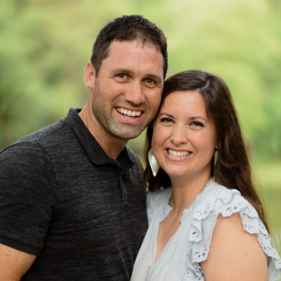A smiling couple standing closely together outdoors with a green, blurred background.