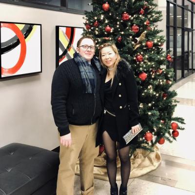 A man and woman in coats smiling together in front of a Christmas tree in a lobby of a building.