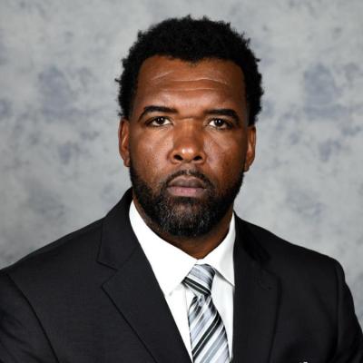A headshot of a man in front of grey background in a black suit.