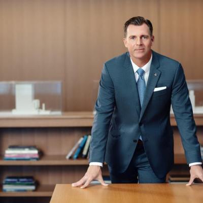 A professionally dressed man in a suit leans forward on a desk in a modern office setting.