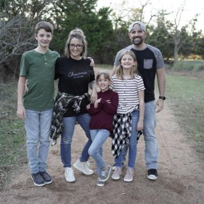 A family of five posing outdoors on a dirt path, surrounded by trees.