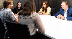 Six professionals sit around a white conference table, engaged in a group discussion in a modern office setting.