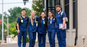 A group of five nursing students in blue scrubs, standing outside a building, smiling at the camera with stethoscopes around their necks.