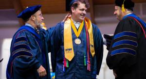 Two professors in academic regalia place a stole around a male graduate on a stage.