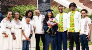 A graduate in a navy cap and gown poses with her family dressed in traditional attire in front of the East Texas Baptist University historical marker.