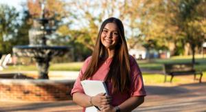 A young woman with brown hair and a pink shirt holding a notebook standing infront of a fountain