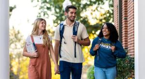 Three young adults walk together outdoors on a bright day, smiling and conversing in front of a brick building with a white column, surrounded by lush greenery.